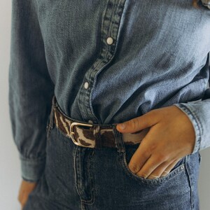 Pode incluir: Uma camisa de ganga com gola com folhos e bot&otilde;es &eacute; combinada com cal&ccedil;as de ganga escuras. Um cinto com padr&atilde;o castanho e branco com fivela dourada aperta a cintura. A m&atilde;o da pessoa est&aacute; no bolso das cal&ccedil;as de ganga.