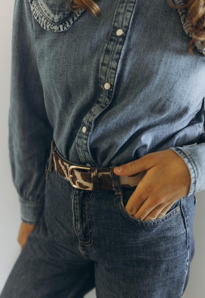 Pode incluir: Uma camisa de ganga com gola com folhos e bot&otilde;es &eacute; combinada com cal&ccedil;as de ganga escuras. Um cinto com padr&atilde;o castanho e branco com fivela dourada aperta a cintura. A m&atilde;o da pessoa est&aacute; no bolso das cal&ccedil;as de ganga.