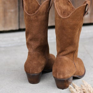 K&ouml;nnte beinhalten: Ein Paar braune Wildleder-Cowbo Stiefel mit niedrigem Absatz. Die Stiefel stehen auf einer Betonfl&auml;che.