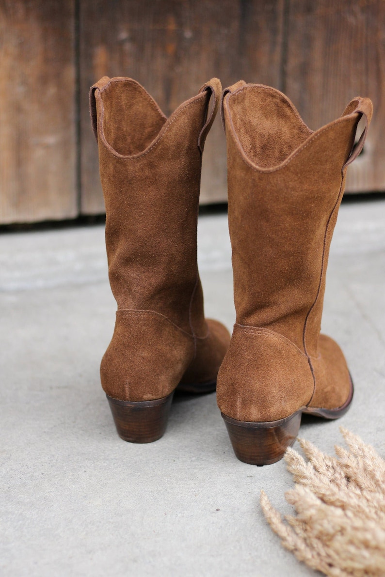 K&ouml;nnte beinhalten: Ein Paar braune Wildleder-Cowbo Stiefel mit niedrigem Absatz. Die Stiefel stehen auf einer Betonfl&auml;che.
