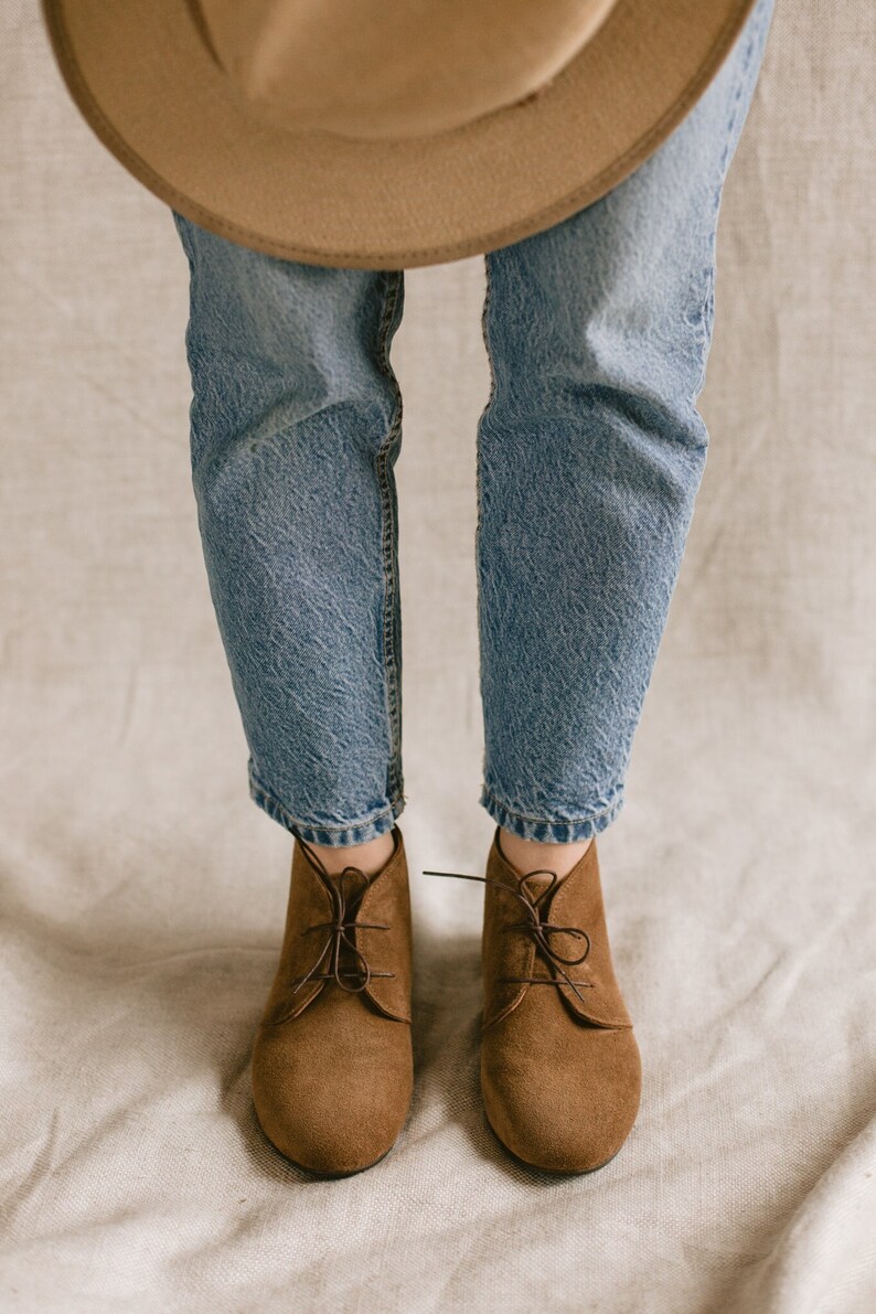 May include: A pair of brown suede ankle boots with laces. The boots are worn with blue jeans and a brown hat.