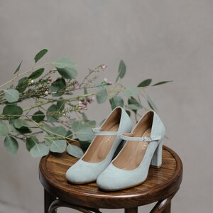 May include: A pair of light blue suede heeled shoes with ankle straps on a wooden stool. The shoes have a block heel and a rounded toe.