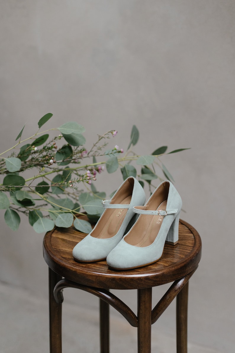 May include: A pair of light blue suede heeled shoes with ankle straps on a wooden stool. The shoes have a block heel and a rounded toe.