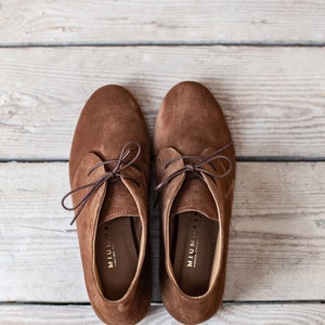 May include: A pair of brown suede lace-up shoes with a low heel. The shoes have a simple design and are made of soft, comfortable material. The brand name "MIUMIA" is printed on the inside of each shoe.