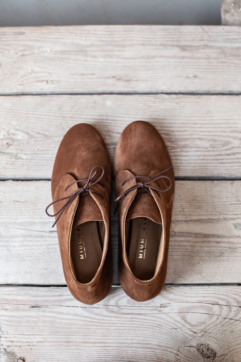 May include: A pair of brown suede lace-up shoes with a low heel. The shoes have a simple design and are made of soft, comfortable material. The brand name "MIUMIA" is printed on the inside of each shoe.