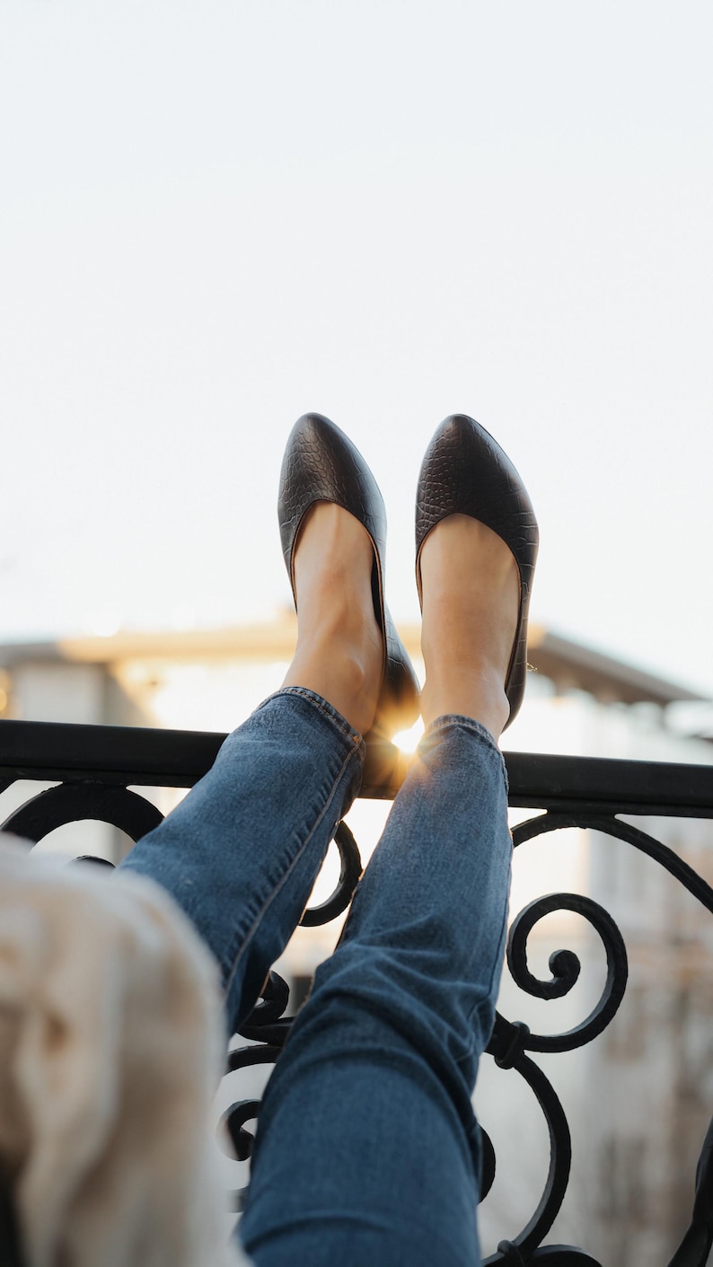 May include: A pair of dark brown leather flats with a textured finish. The shoes are worn with blue jeans, with the feet resting on a black metal railing. The sun shines brightly in the background.