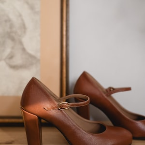 May include: A pair of brown leather heeled shoes with a strap and buckle. The shoes are on a wooden surface.