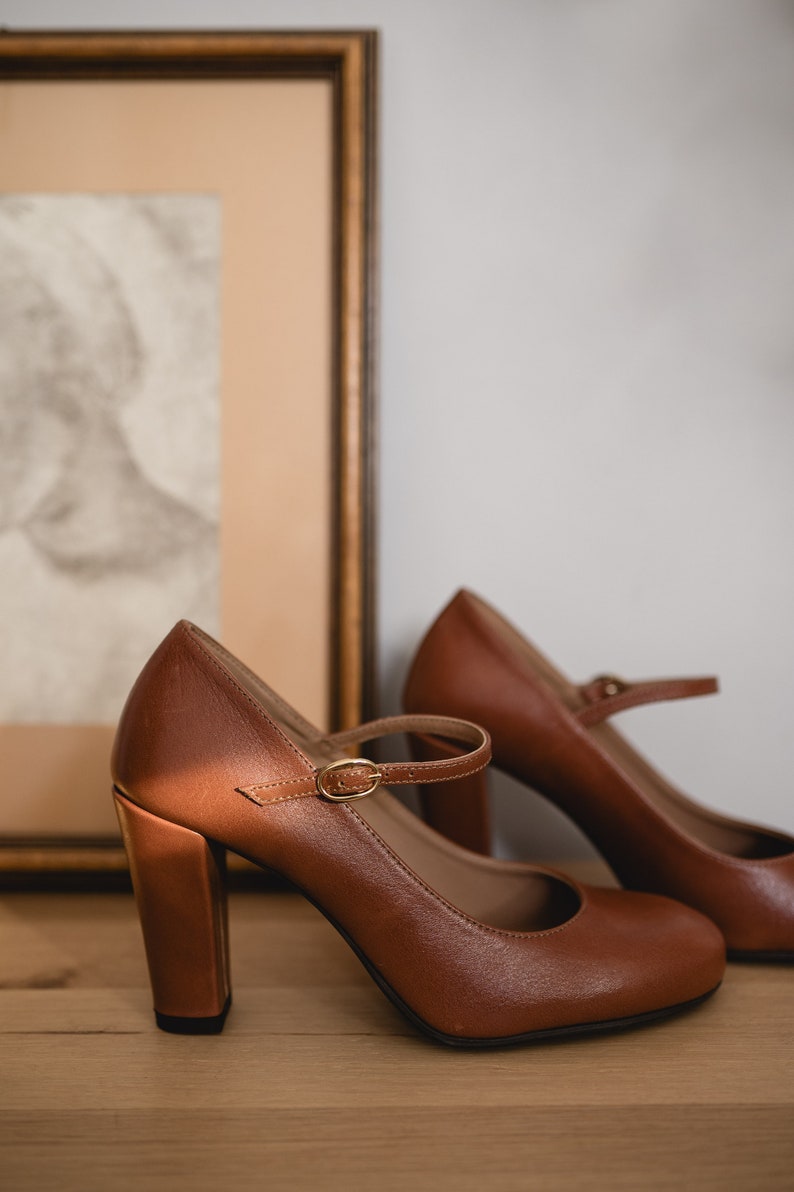 May include: A pair of brown leather heeled shoes with a strap and buckle. The shoes are on a wooden surface.