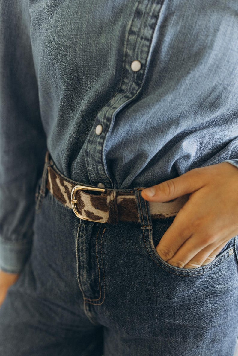 Pode incluir: Um cinto de pele de vaca castanho e branco com fivela dourada &eacute; usado com uma camisa de ganga e cal&ccedil;as de ganga. O cinto &eacute; o ponto focal, adicionando textura e padr&atilde;o. A fivela &eacute; retangular.