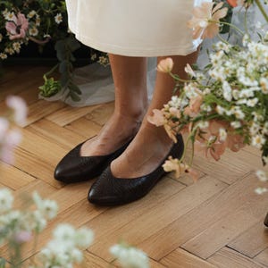 May include: A pair of dark brown leather flats with a textured surface. The shoes have a pointed toe design. The image is taken from a low angle, showing the shoes and the lower part of a white dress. The background includes flowers and wooden flooring.