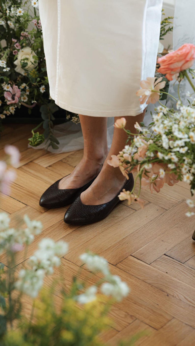May include: A pair of dark brown leather flats with a textured surface. The shoes have a pointed toe design. The image is taken from a low angle, showing the shoes and the lower part of a white dress. The background includes flowers and wooden flooring.