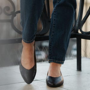 May include: Close-up of black leather ballet flats with a textured finish. The shoes are worn with dark blue denim jeans. The image is taken from a low angle, highlighting the footwear.