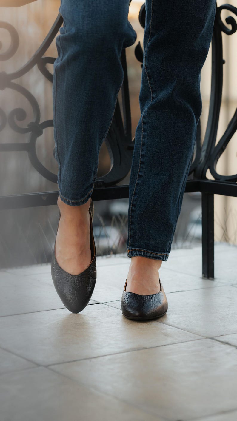 May include: Close-up of black leather ballet flats with a textured finish. The shoes are worn with dark blue denim jeans. The image is taken from a low angle, highlighting the footwear.