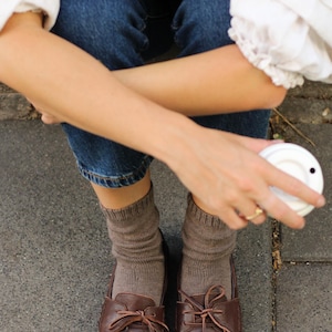 Puede incluir: Un par de mocasines de cuero marr&oacute;n oscuro con cordones finos atados en lazos en la parte superior, usados con calcetines acanalados de color topo. El calzado presenta costuras visibles a lo largo de la punta redondeada y un acabado mate.