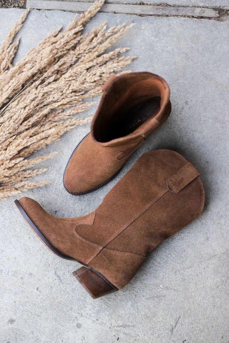 K&ouml;nnte beinhalten: Ein Paar braune Wildleder-Cowbo Stiefel mit spitzer Zehenpartie und Blockabsatz. Die Stiefel liegen auf einer grauen Betonfl&auml;che mit einem B&uuml;ndel getrockneten Grases im Hintergrund.