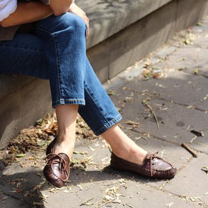 Puede incluir: Un par de mocasines de cuero marr&oacute;n oscuro con punta redondeada, detalles de costuras visibles a lo largo de los bordes y un cord&oacute;n de cuero fino atado en un lazo sobre el empeine. Los zapatos se llevan puestos con pantalones vaqueros azules.
