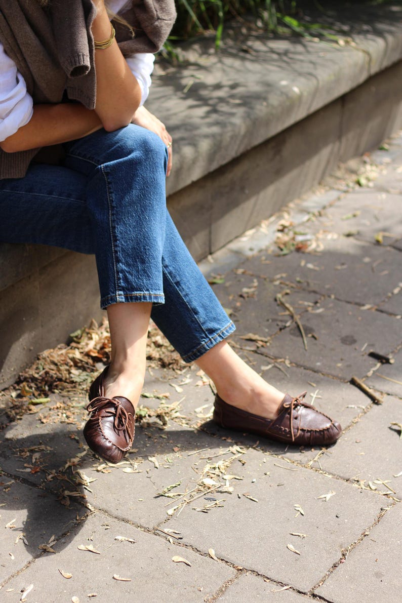 Puede incluir: Un par de mocasines de cuero marr&oacute;n oscuro con punta redondeada, detalles de costuras visibles a lo largo de los bordes y un cord&oacute;n de cuero fino atado en un lazo sobre el empeine. Los zapatos se llevan puestos con pantalones vaqueros azules.