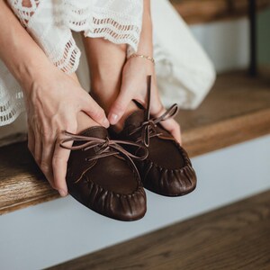 Puede incluir: Un par de mocasines de cuero marr&oacute;n oscuro con costura fruncida en la puntera, puntera redondeada y cordones finos de cuero a juego atados en un lazo en el centro del empeine. Los zapatos tienen una construcci&oacute;n suave y flexible.