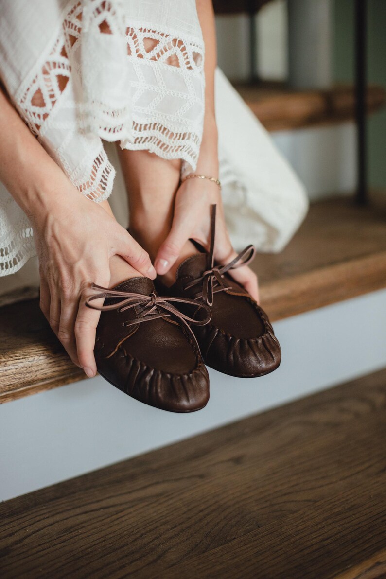 Puede incluir: Un par de mocasines de cuero marr&oacute;n oscuro con costura fruncida en la puntera, puntera redondeada y cordones finos de cuero a juego atados en un lazo en el centro del empeine. Los zapatos tienen una construcci&oacute;n suave y flexible.