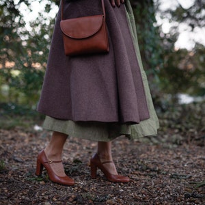 May include: A woman wearing a brown coat and a brown leather bag over her shoulder. She is wearing a brown skirt and brown shoes with a buckle.
