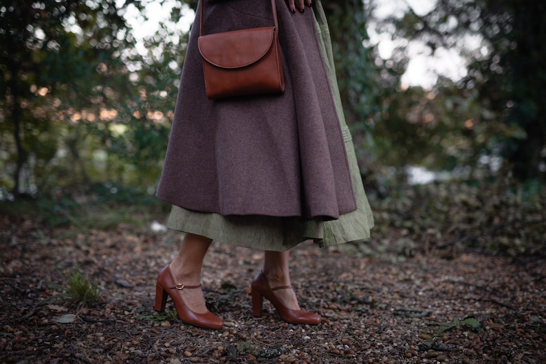 May include: A woman wearing a brown coat and a brown leather bag over her shoulder. She is wearing a brown skirt and brown shoes with a buckle.