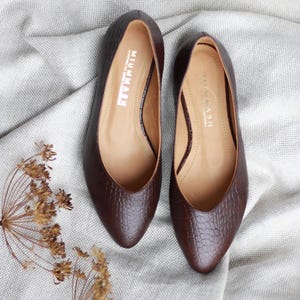 May include: A pair of brown leather flats with a pointed toe and a textured, crocodile-like pattern. The shoes have a low heel and the brand name "MIUMMASH" is visible inside. The flats are displayed on a light gray fabric.