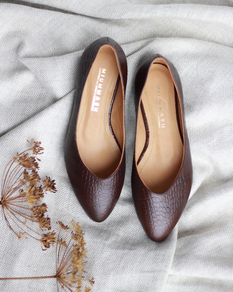 May include: A pair of brown leather flats with a pointed toe and a textured, crocodile-like pattern. The shoes have a low heel and the brand name "MIUMMASH" is visible inside. The flats are displayed on a light gray fabric.