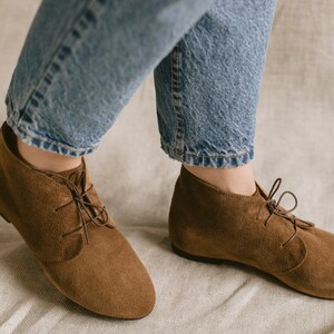 May include: A pair of brown suede ankle boots with laces. The boots are worn by a person wearing blue jeans.