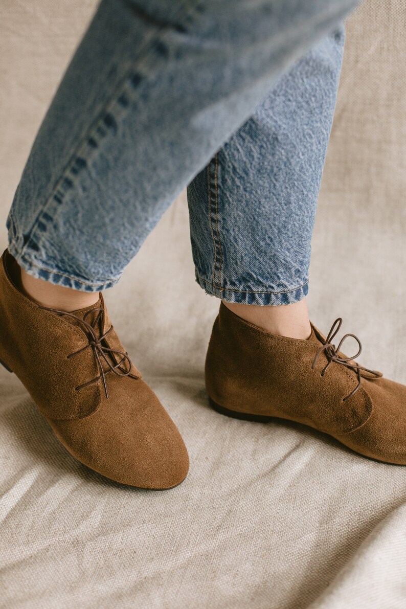 May include: A pair of brown suede ankle boots with laces. The boots are worn by a person wearing blue jeans.