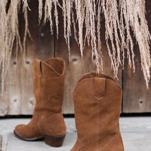 K&ouml;nnte beinhalten: Ein Paar braune Wildleder-Cowbo Stiefel mit spitzer Zehenpartie und niedrigem Absatz. Die Stiefel stehen auf einer Betonfl&auml;che vor einer Holzwand, an der getrocknetes Gras h&auml;ngt.