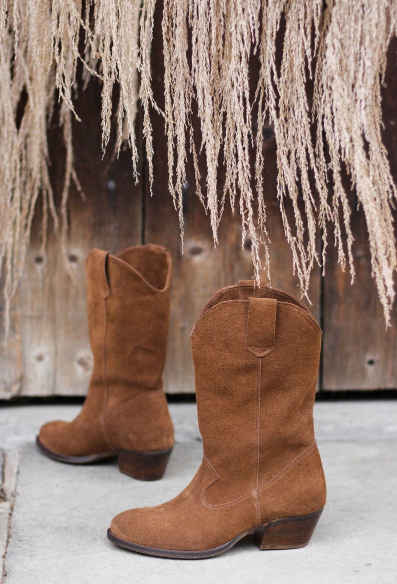 K&ouml;nnte beinhalten: Ein Paar braune Wildleder-Cowbo Stiefel mit spitzer Zehenpartie und niedrigem Absatz. Die Stiefel stehen auf einer Betonfl&auml;che vor einer Holzwand, an der getrocknetes Gras h&auml;ngt.