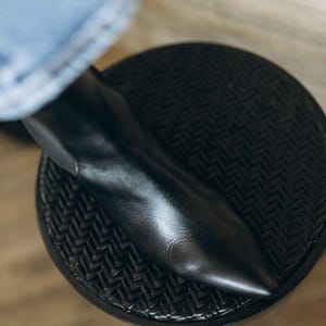 Puede incluir: Un bot&iacute;n negro de cuero con punta afilada, acabado liso y silueta limpia. El bot&iacute;n est&aacute; colocado sobre un taburete circular oscuro con una superficie superior que presenta un patr&oacute;n de tejido en espiga.