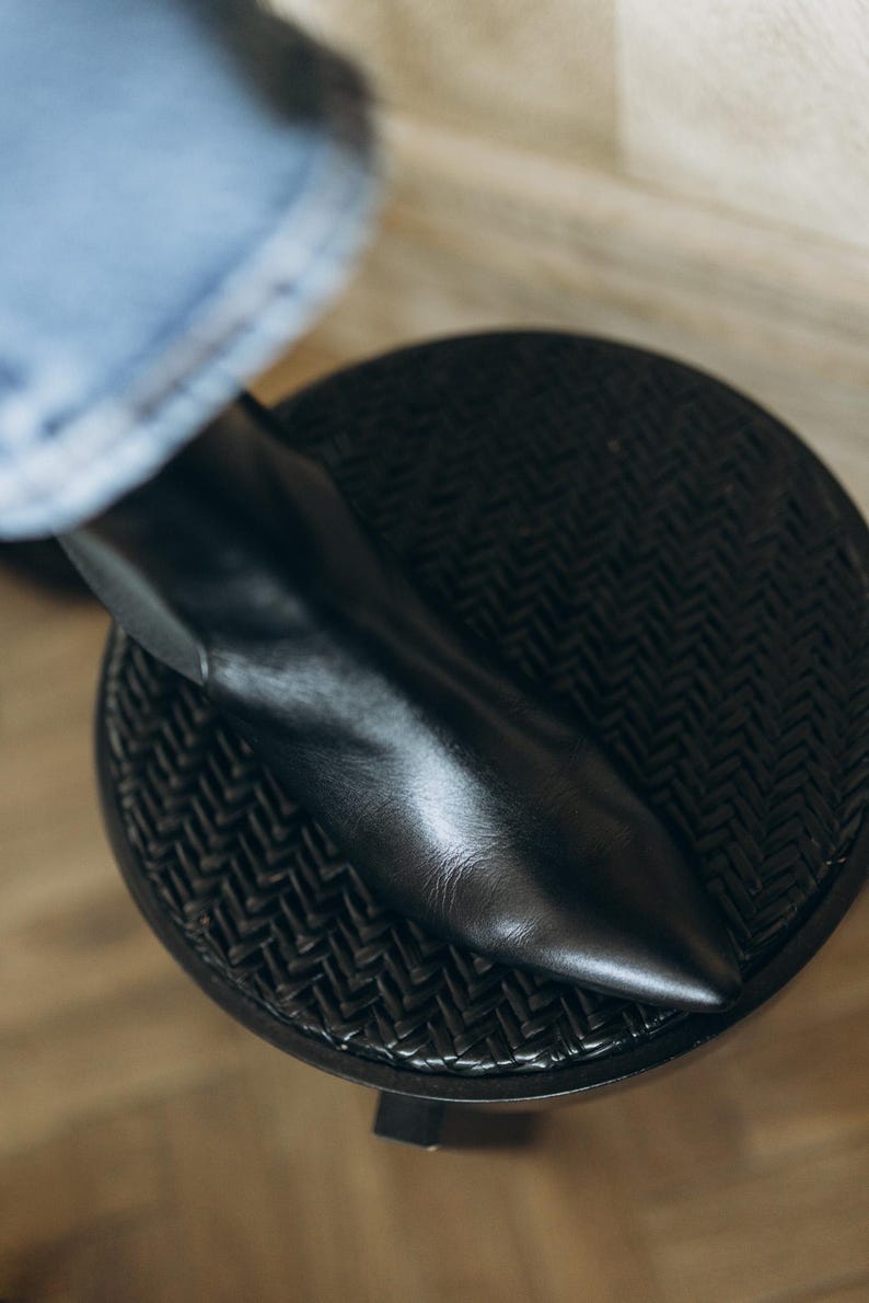 Puede incluir: Un bot&iacute;n negro de cuero con punta afilada, acabado liso y silueta limpia. El bot&iacute;n est&aacute; colocado sobre un taburete circular oscuro con una superficie superior que presenta un patr&oacute;n de tejido en espiga.