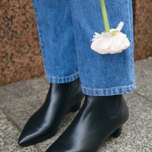 Puede incluir: Un par de botines de cuero negro con punta afilada y tac&oacute;n peque&ntilde;o, usados con pantalones vaqueros azules. Una flor de color rosa p&aacute;lido con un tallo verde largo est&aacute; colocada frente a la tela de mezclilla azul de los pantalones.