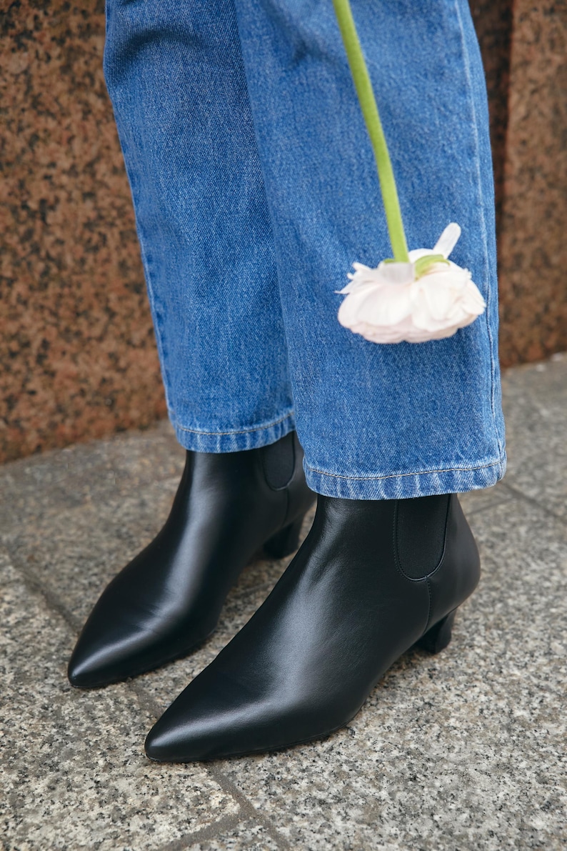Puede incluir: Un par de botines de cuero negro con punta afilada y tac&oacute;n peque&ntilde;o, usados con pantalones vaqueros azules. Una flor de color rosa p&aacute;lido con un tallo verde largo est&aacute; colocada frente a la tela de mezclilla azul de los pantalones.