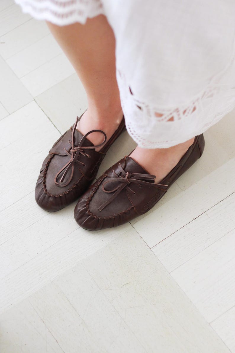 Puede incluir: Un par de mocasines de cuero marr&oacute;n oscuro con costura fruncida en la punta, un lazo decorativo de cuero en la parte superior y una silueta de perfil bajo. Los zapatos presentan costuras visibles a lo largo del borde de la puntera.
