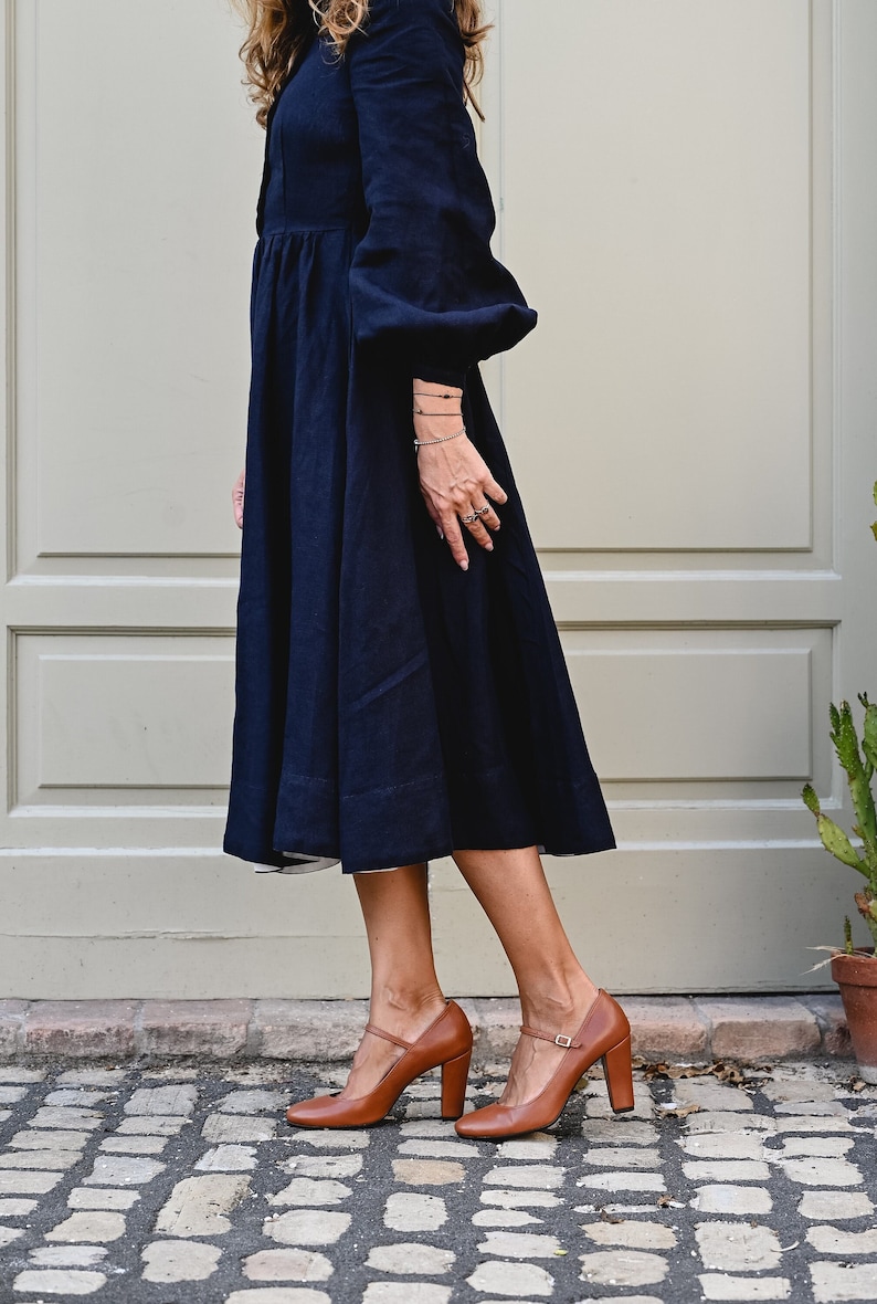May include: A woman wearing a navy blue midi dress with long sleeves and a gathered skirt. She is wearing brown leather heeled shoes with a buckle strap.