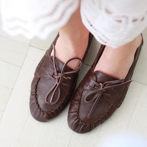 Puede incluir: Un par de mocasines de cuero marr&oacute;n oscuro con punta redondeada, una costura fruncida a lo largo del borde delantero y un cord&oacute;n de cuero decorativo atado en un lazo en la parte superior. Los zapatos tienen un acabado liso y perfil bajo.