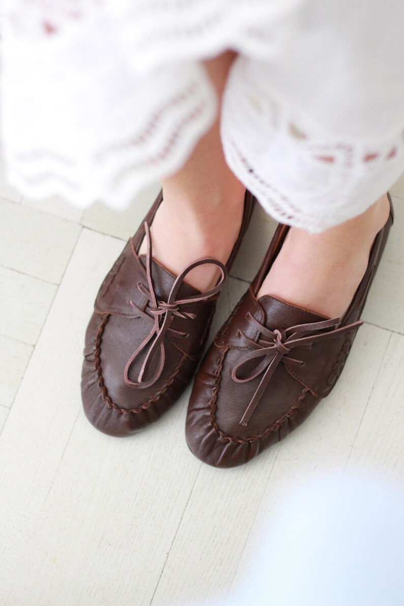 Puede incluir: Un par de mocasines de cuero marr&oacute;n oscuro con punta redondeada, una costura fruncida a lo largo del borde delantero y un cord&oacute;n de cuero decorativo atado en un lazo en la parte superior. Los zapatos tienen un acabado liso y perfil bajo.