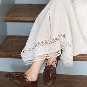 Puede incluir: Un par de zapatos de cuero marr&oacute;n oscuro estilo mocas&iacute;n con cordones finos y costura fruncida en la punta, combinados con una falda larga de tela texturizada color blanco roto con ribete de encaje blanco en el dobladillo.