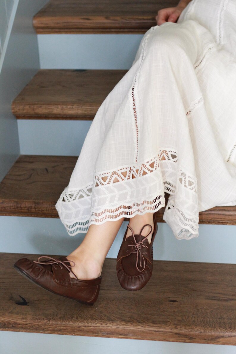 Puede incluir: Un par de zapatos de cuero marr&oacute;n oscuro estilo mocas&iacute;n con cordones finos y costura fruncida en la punta, combinados con una falda larga de tela texturizada color blanco roto con ribete de encaje blanco en el dobladillo.