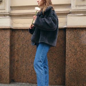 Puede incluir: Una boina rosa claro, una chaqueta oversize de piel sint&eacute;tica gris oscuro con cuello ancho, pantalones vaqueros azules y botines negros de punta fina. El conjunto se completa con una flor de color rosa p&aacute;lido sostenida en la mano.