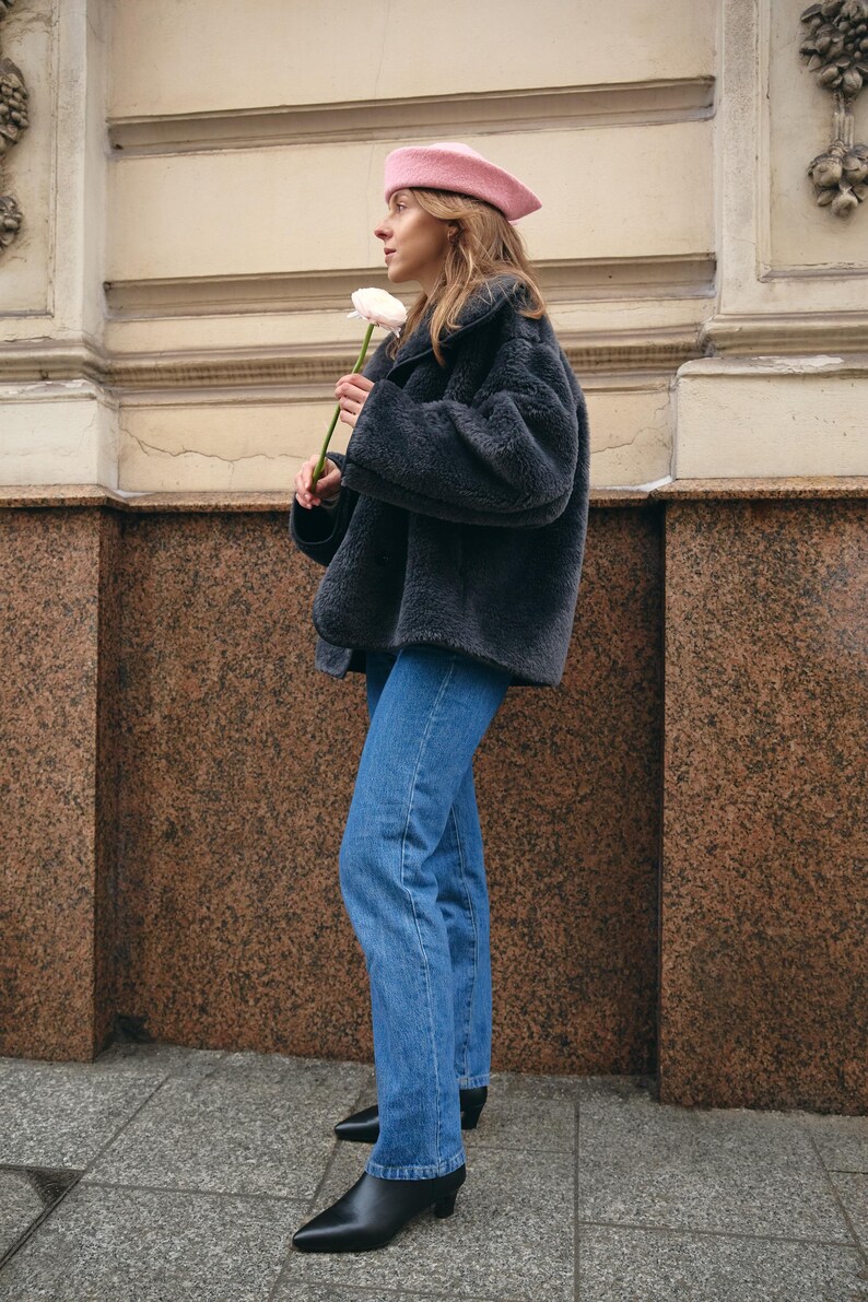 Puede incluir: Una boina rosa claro, una chaqueta oversize de piel sint&eacute;tica gris oscuro con cuello ancho, pantalones vaqueros azules y botines negros de punta fina. El conjunto se completa con una flor de color rosa p&aacute;lido sostenida en la mano.