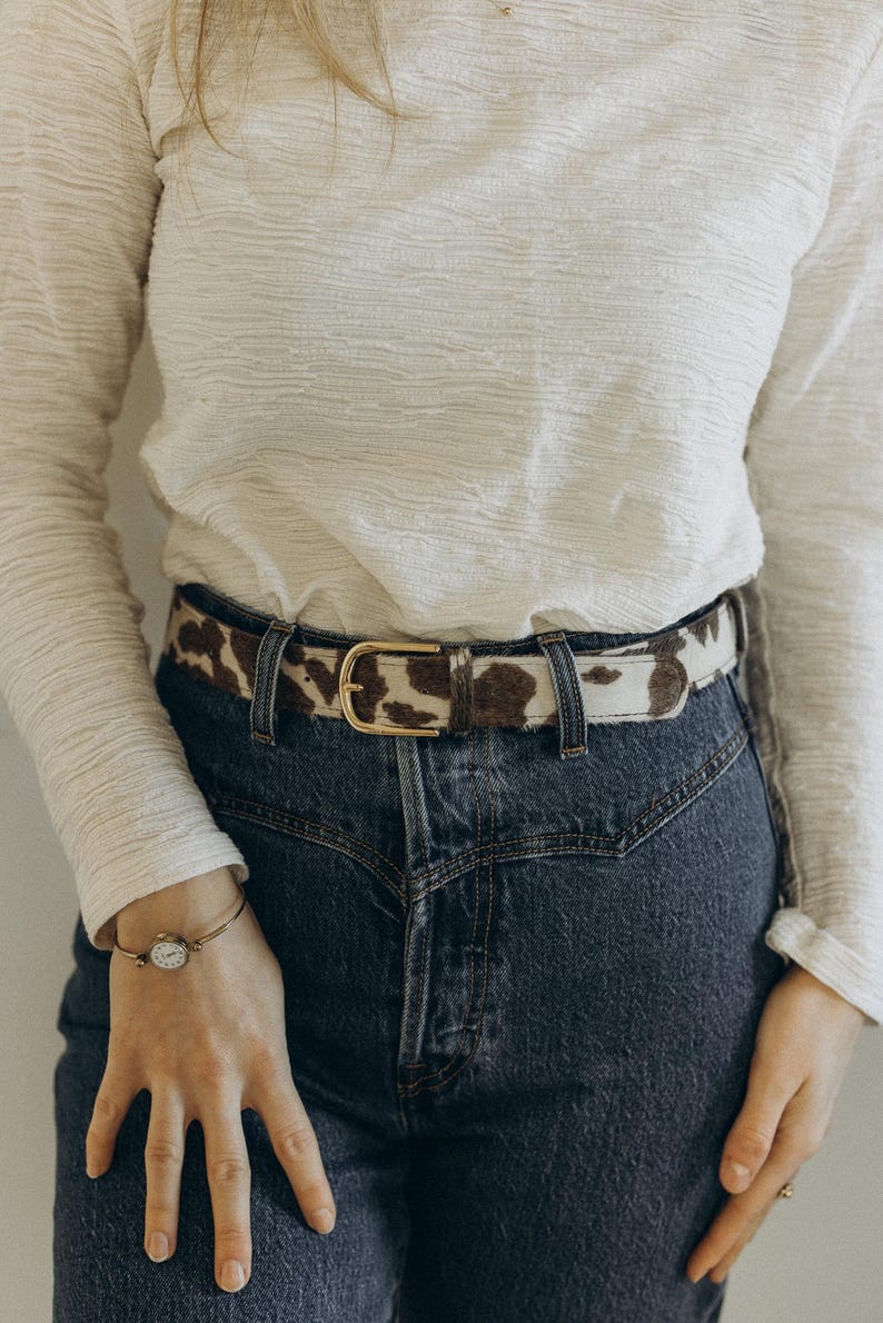Pode incluir: Um cinto de pele de vaca com fivela dourada &eacute; usado com jeans azuis. O cinto apresenta um padr&atilde;o de manchas marrons e brancas. A pessoa tamb&eacute;m est&aacute; usando uma camisa branca de manga comprida e uma pulseira dourada.