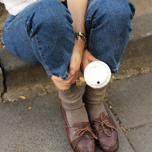 Puede incluir: Una camisa blanca de manga larga, pantalones vaqueros azules, calcetines de punto marrones y mocasines de cuero marr&oacute;n oscuro con cordones. Una pulsera de metal dorado en la mu&ntilde;eca y un vaso de caf&eacute; desechable blanco sostenido en la mano.