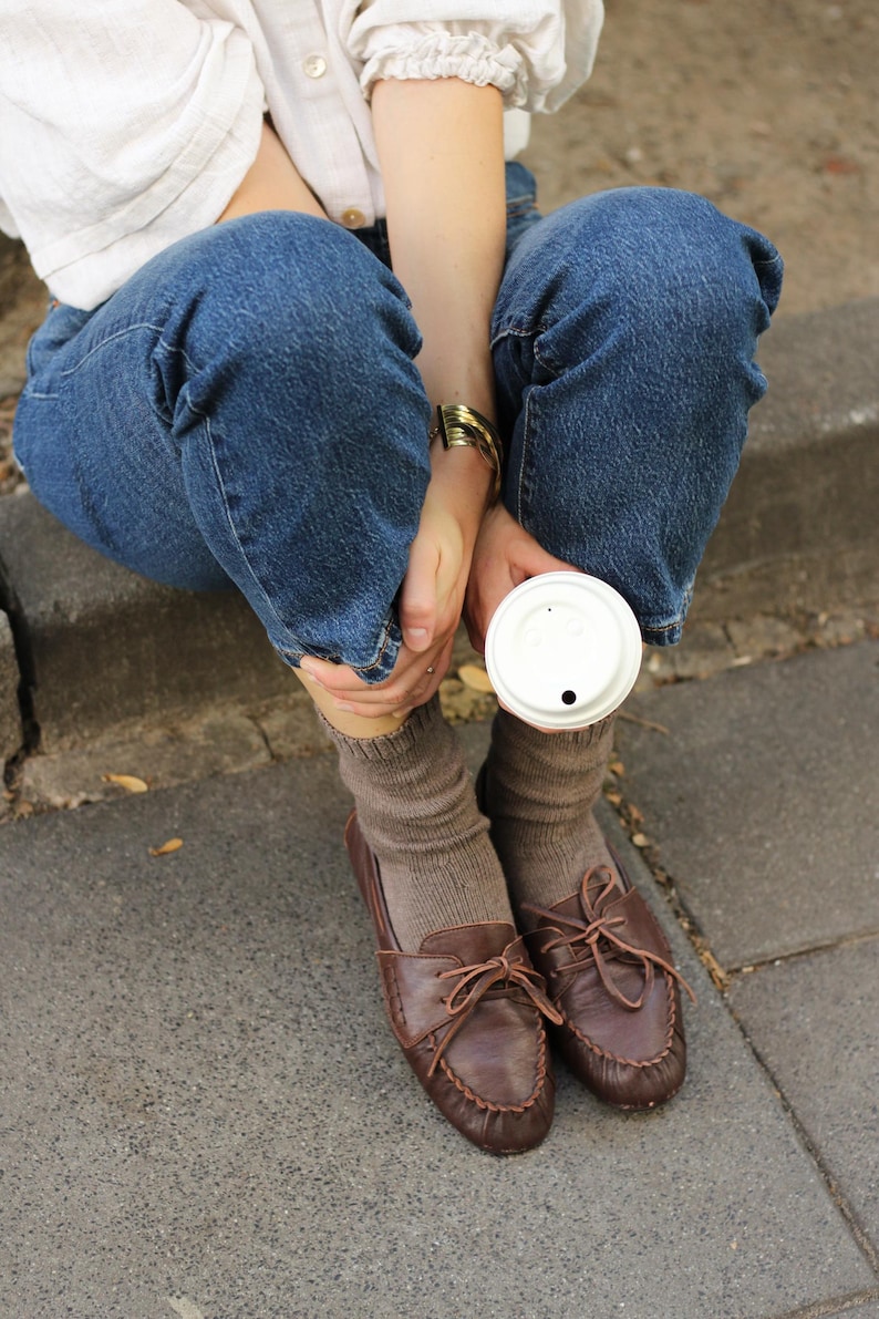 Puede incluir: Una camisa blanca de manga larga, pantalones vaqueros azules, calcetines de punto marrones y mocasines de cuero marr&oacute;n oscuro con cordones. Una pulsera de metal dorado en la mu&ntilde;eca y un vaso de caf&eacute; desechable blanco sostenido en la mano.
