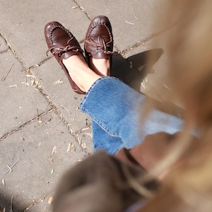 Puede incluir: Un par de mocasines de cuero marr&oacute;n oscuro con punta redondeada, una costura decorativa a lo largo de la parte delantera y un cord&oacute;n de cuero fino atado en un lazo en la parte superior. Los zapatos se llevan con pantalones vaqueros azules.