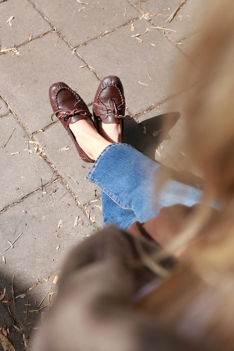 Puede incluir: Un par de mocasines de cuero marr&oacute;n oscuro con punta redondeada, una costura decorativa a lo largo de la parte delantera y un cord&oacute;n de cuero fino atado en un lazo en la parte superior. Los zapatos se llevan con pantalones vaqueros azules.