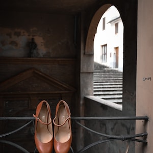 May include: A pair of brown leather shoes with a strap and buckle detail are hanging on a black metal railing. The shoes are in front of a window with a view of a stone staircase.