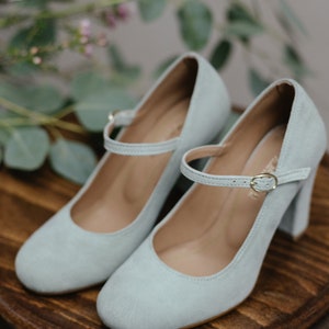 May include: A pair of light blue suede heeled shoes with a buckle strap. The shoes have a block heel and a rounded toe.